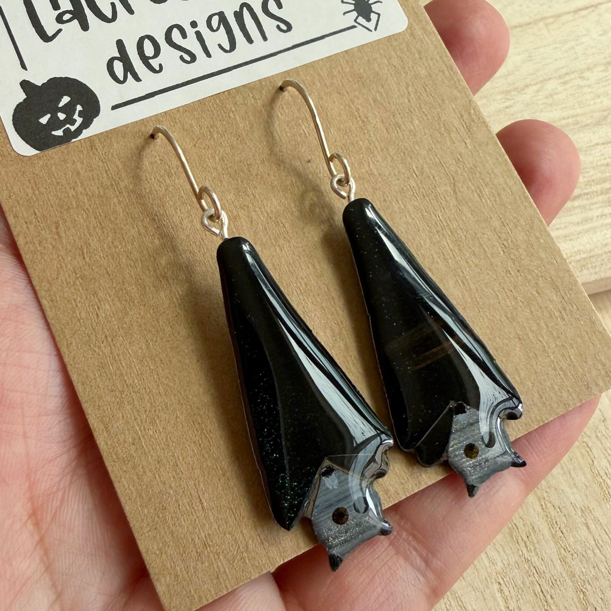 Black bat-shaped drop earrings with silver hooks, displayed on a brown kraft card labeled 'Lacroz & Co. designs' held in a hand.